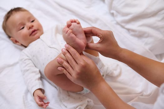 Close-up of little feet with tiny toes in the hands of a mother of blurred adorable baby boy smiling looking at his mom cuddling him. Babyhood and infancy. Maternity. Motherhood and baby care concept