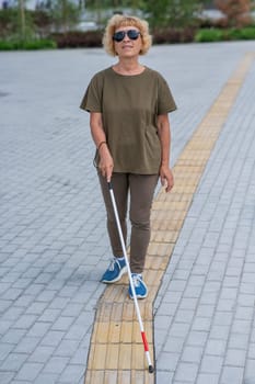 An elderly blind woman walks with a cane along a tactile tile