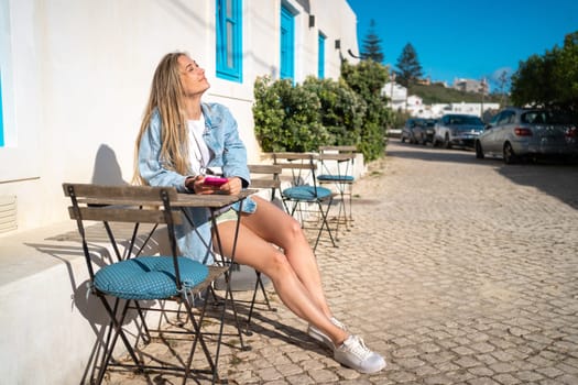 Woman sitting outdoor European cafe holding smartphone in hand enjoy summer vacation in typical portugal city