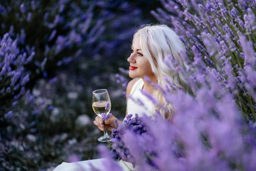 Blonde lavender field holds a glass of white wine in her hands. Happy woman in white dress enjoys lavender field picnic holding a large bouquet of lavender in her hands . Illustrating woman's picnic in a lavender field