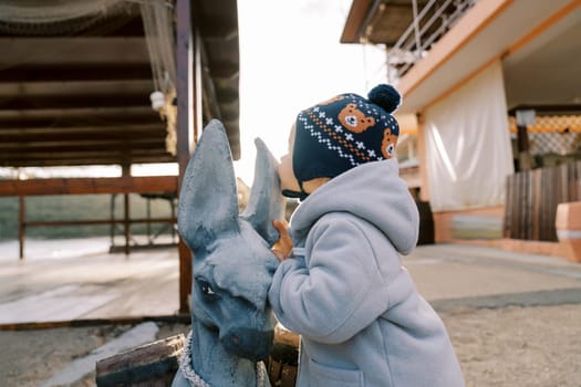 Little girl kisses the ear of a statue of a fabulous donkey loaded with barrels. High quality photo