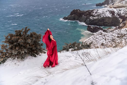 Woman red dress snow sea. Happy woman in a red dress in the snowy mountains by the emerald sea. The wind blows her clothes, posing against sea and snow background