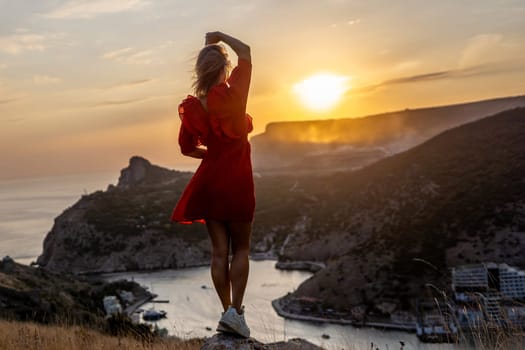 Happy woman standing with her back on the sunset in nature in summer with open hands posing with mountains on sunset, silhouette. Woman in the mountains red dress, eco friendly, summer landscape active rest.