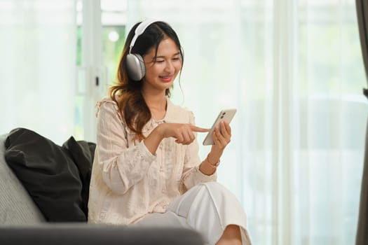 Pretty young Asian woman in headphones watching streaming tv videos online on smartphone.