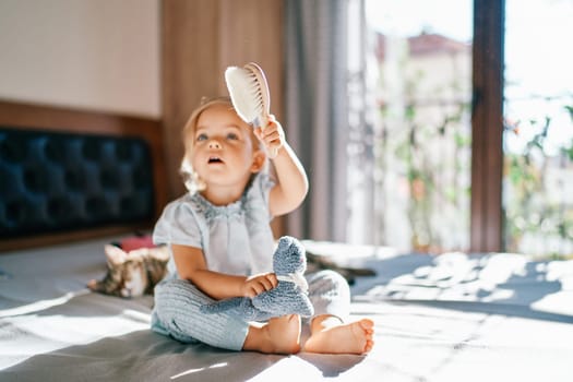 Little girl sits on a bed with a toy on her knees and holds out a hairbrush. High quality photo