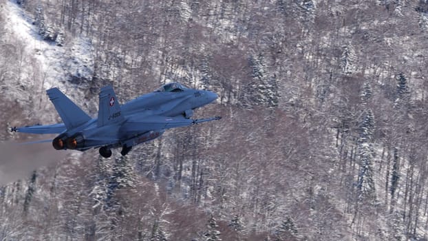 Meiringen Switzerland January 19 2023: Fighter jet ascends powerfully with its afterburners flaming against the serene backdrop of a narrow, snow-covered Alpine valley. Copy Space.