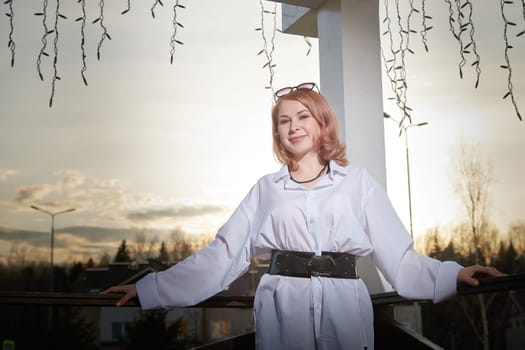 Portrait of Beautiful girl with red hair in white shirt in open wooden pavillion in village or small town. Young slender woman and sky with clouds on background on autumn, spring or summer evening