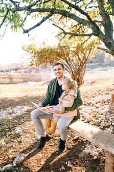 Smiling dad with a little girl on his lap sitting on a bench with a colorful book in his hands. High quality photo