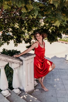 woman in a red silk dress and a bandage on her head smiles against the background of the leaves of a tree. She is leaning on the coop and looking into the camera. Vertical photo