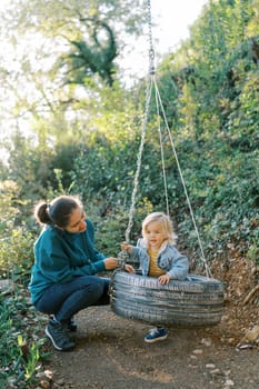 Mom is squatting next to a little girl sitting on a tire swing in the park. High quality photo