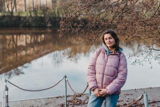 A young woman standing at the shore looking at the river in autumn sunny day. Street view, copy space for text, travel photo. Happy tourist woman on the bank of the river in autumn in warm clothes. Tourists enjoy their vacation, winter season. Romantic look and travel concept. A joyful mood in a Caucasian girl. Winter Wonderland: Enchanting Girl by the Riverside in Autumn.