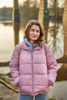 A young woman standing at the shore looking at the river in autumn sunny day. Street view, copy space for text, travel photo. Happy tourist woman on the bank of the river in autumn in warm clothes. Tourists enjoy their vacation, winter season. Romantic look and travel concept. A joyful mood in a Caucasian girl. Winter Wonderland: Enchanting Girl by the Riverside in Autumn.