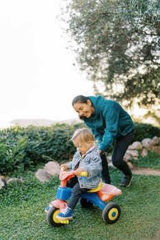 Mom pushes a little girl riding a tricycle on a green lawn. High quality photo