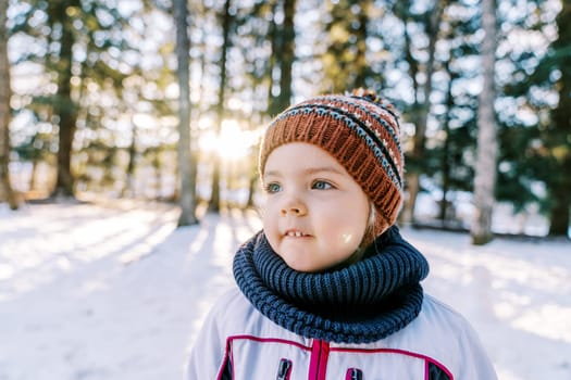 Little smiling girl in a hat and scarf stands in a snowy forest and looks away. High quality photo