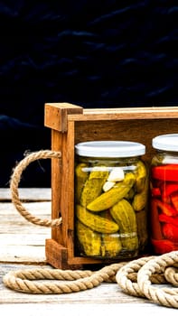 Wooden crate with glass jars with pickled red bell peppers and pickled cucumbers (pickles) isolated. Jars with variety of pickled vegetables. Preserved food concept in a rustic composition.