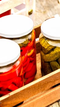 Wooden crate with glass jars with pickled red bell peppers and pickled cucumbers (pickles) isolated. Jars with variety of pickled vegetables. Preserved food concept in a rustic composition.