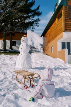 Little child sits in the snow near a sled next to a snowman in the courtyard of a wooden house. Back view. High quality photo