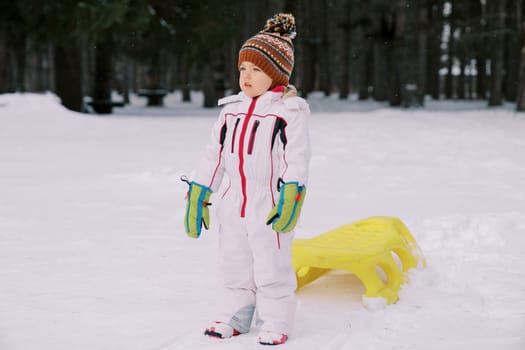 Little girl stands sideways in the snow near a yellow sled at the edge of the forest. High quality photo