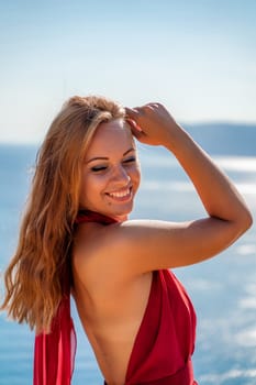 Smiling young woman in a red dress looks at the camera. A beautiful tanned girl enjoys her summer holidays at the sea. Portrait of a stylish carefree woman laughing at the ocean