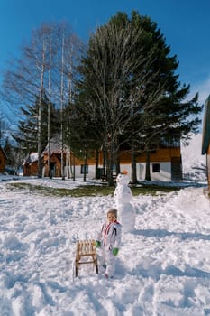Little girl stands near a sled next to a snowman in the yard of a wooden house. High quality photo