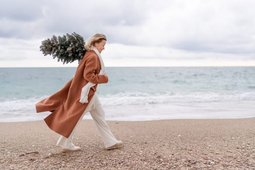 Blond woman Christmas sea. Christmas portrait of a happy woman walking along the beach and holding a Christmas tree on her shoulder. She is wearing a brown coat and a white suit