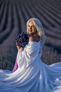 Blonde woman poses in lavender field at sunset. Happy woman in white dress holds lavender bouquet. Aromatherapy concept, lavender oil, photo session in lavender.