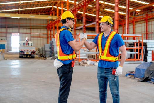 Two Caucasian technician workers hold hands and look happy in factory workplace area with backgroud of truck and the products.