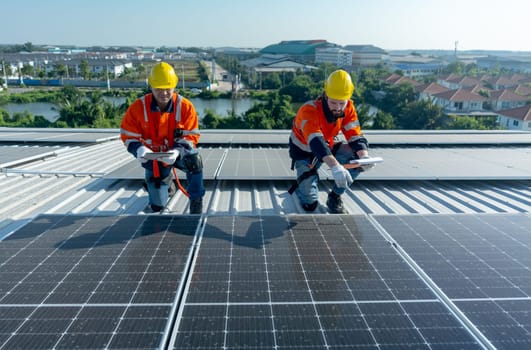 Two Caucasian technician workers hold document pad and tablet discuss about the maintenance of solar cell panels on rooftop of the building or factory.