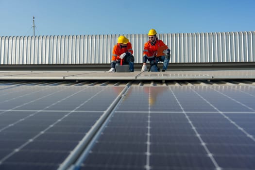 Two Caucasian technician workers sit on rooftop and discuss about the maintenance solar cell panels on rooftop of factory or the building. Green and sustainable energy concept.