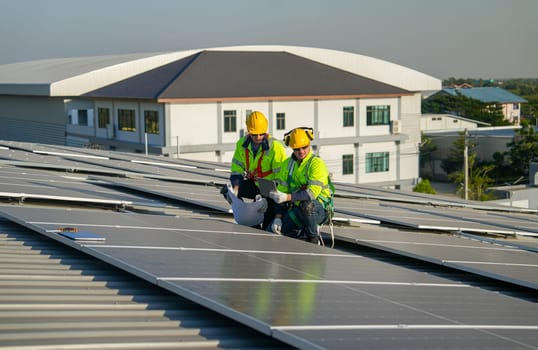 Two Caucasian technician workers use construction plan and tablet to check and maintenance solar cell panels on rooftop of factory or the building. Green and sustainable energy for good environment.