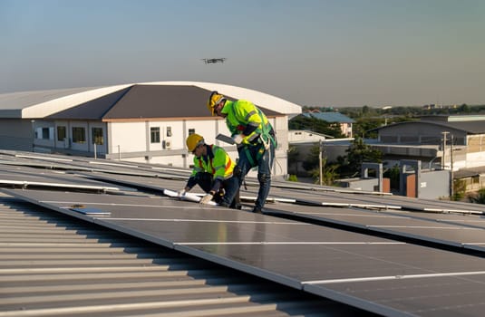 One of technician worker look at construction plan to maintenance solar cell panels and other co-worker hold tablet to work together on rooftop of factory building.