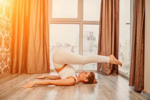 Young athletic attractive woman practicing yoga. Works out at home or in a yoga studio, sportswear, white pants and a full-length top indoors. Healthy lifestyle concept.