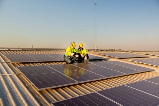 Wide shot two Caucasian technician workers sit on rooftop and discuss about the maintenance solar cell panels on rooftop of factory or the building. Green and sustainable energy concept.