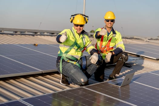 Two industrial technician worker sit near solar cell panels on rooftop of factory building and show thumbs up to camera with smiling. Green and sustainable energy concept.