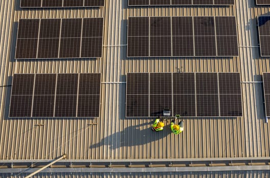 Top view or drone view of two technician workers use laptop and tablet to check and maintenance solar cell panels on rooftop of factory building with green and sustainable energy concept.