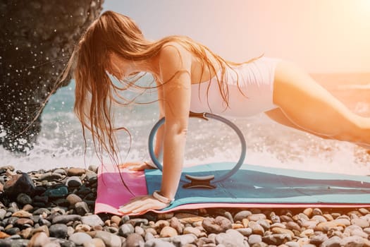 Young woman with black hair, fitness instructor in pink sports leggings and tops, doing pilates on yoga mat with magic pilates ring by the sea on the beach. Female fitness daily yoga concept