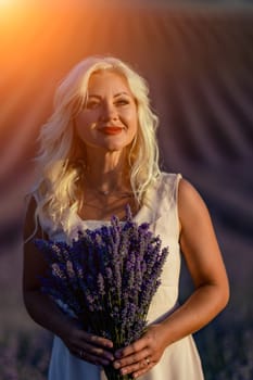 Blonde woman poses in lavender field at sunset. Happy woman in white dress holds lavender bouquet. Aromatherapy concept, lavender oil, photo session in lavender.