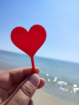 Person holding in fingers hand stick in shape red heart on background sea and sea waves, blue sky in seashore on sunny summer day close-up. Concept love romance amour St Valentines Day