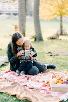 Mom with a little girl in her arms sits on a bedspread in an autumn park and gives her a drink from a mug. High quality photo