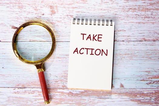 Take Action text written on a notebook on wooden boards next to a magnifying glass