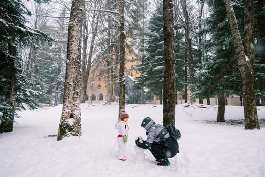 Little girl looks at her mother making a snowman face out of pine cones in the forest. High quality photo