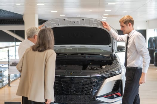 A salesman demonstrates a car with an open hood to an elderly Caucasian couple in a car dealership