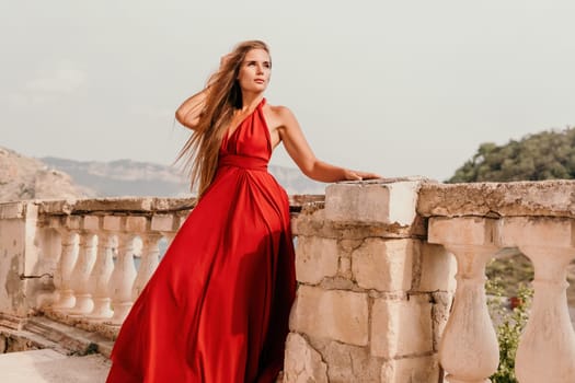 Side view a Young beautiful sensual woman in a red long dress posing on a volcanic rock high above the sea during sunset. Girl on the nature on overcast sky background. Fashion photo