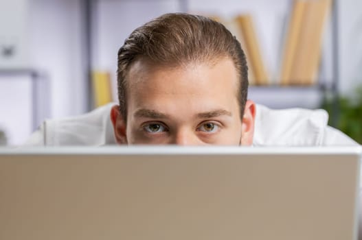 Lebanese confident businessman hiding behind laptop computer, looking at camera, spying his colleagues working, peeping. Professional freelancer man looking from behind computer with cunning eyes