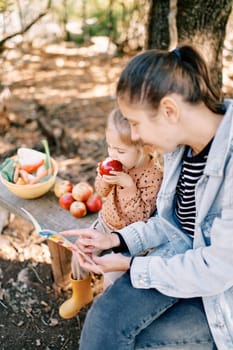 Mom reads a book to a little girl gnawing an apple on a bench in the garden. High quality photo