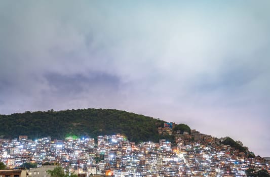 A hillside favela shines at dusk, under a moody sky.