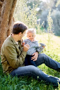 Smiling dad tickling little girl on his knees sitting on the grass. High quality photo