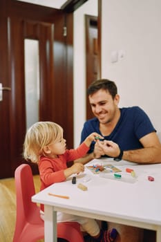 Little girl takes a plasticine figurine from her father hand while sitting at the table. High quality photo