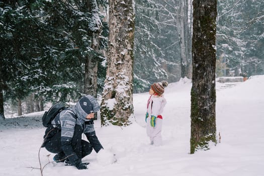 Little girl stands in a snowy forest near her mother making a snowman while squatting. High quality photo