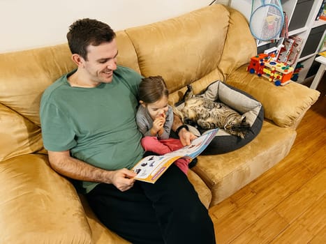 Smiling dad reading book to little girl while sitting on sofa next to cat. High quality photo